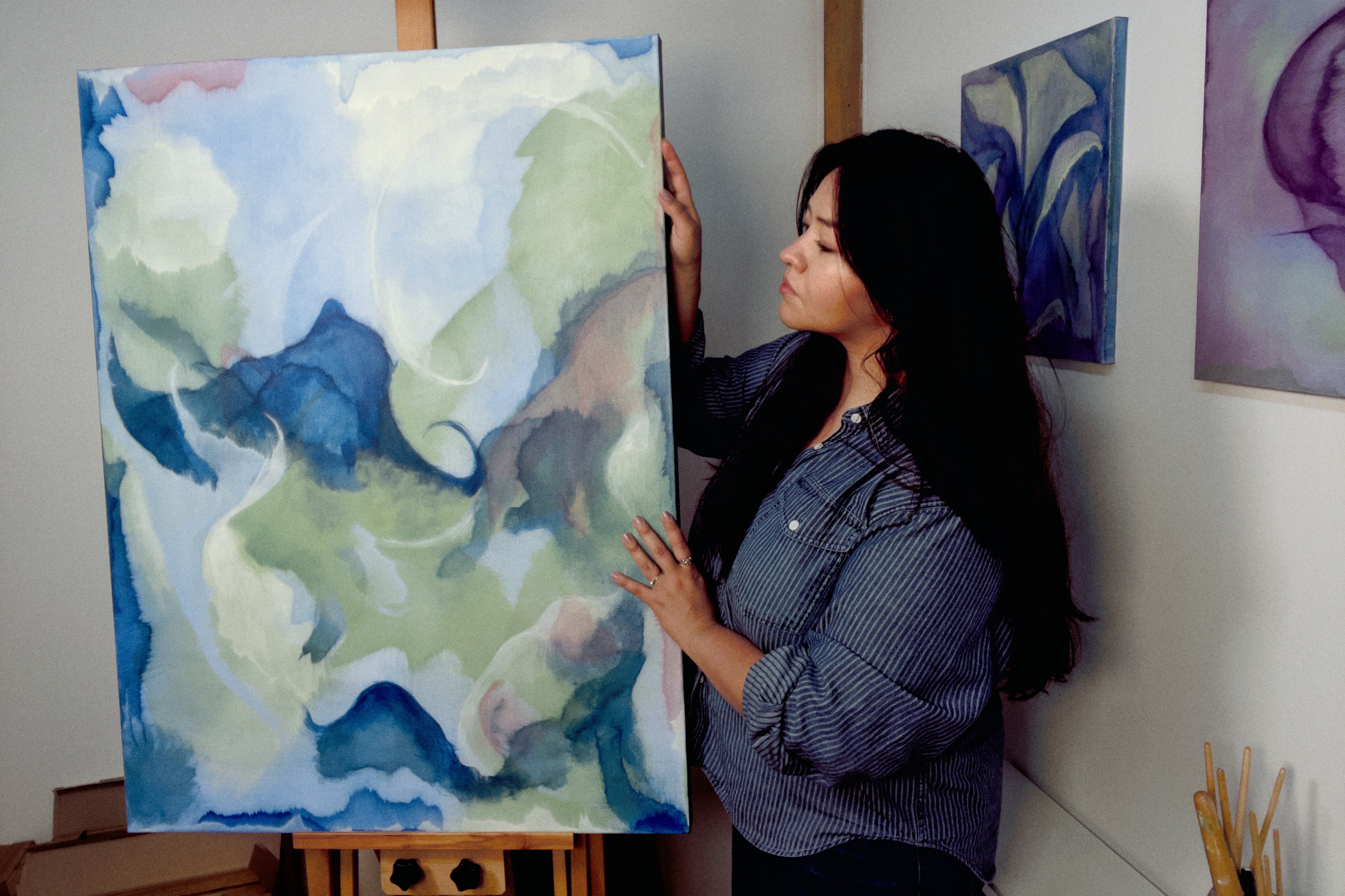 Woman holding a large abstract painting in a room with other artworks on the wall.